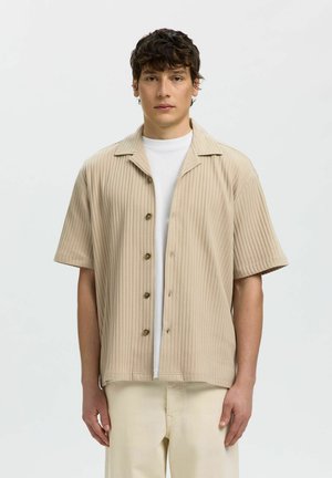 Mladý muž s krátkymi tmavými vlasmi, oblečený v béžovej rebrovanej krátkosrkej košeli na gombíky cez biele tričko a svetlé nohavice.