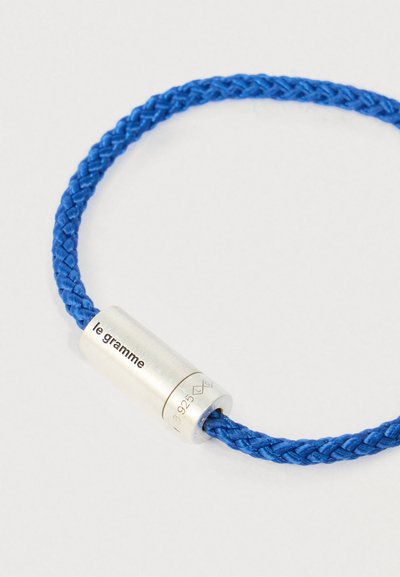 Kék fonott zsinór karkötő ezüst hengeralakú kapoccsal, amelyen a "le gramme" és a "925" minőségi jelzés van gravírozva.