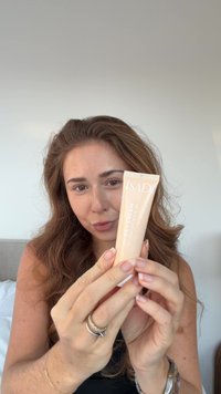 Crème CC dans un tube pêche doux, avec un texte blanc indiquant "ISAD" et "Hydratant SPF 30." Mains tenant le tube sur un fond neutre.