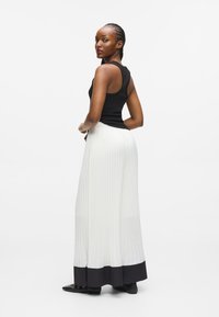 Czarny top bez rękawów z białymi, plisowanymi spodniami w szerokim kroju, z czarnym wykończeniem u dołu. Modelka odwrócona plecami, z warkoczami i czarnym obuwiem.