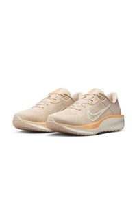 Zapatillas de running beige con parte superior de malla, swoosh blanco de Nike, suela texturizada y acentos en naranja claro en la mediasuela y la lengüeta.