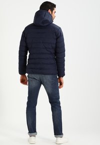 Navy blue puffer jacket with a hood, quilted design, and elastic cuffs. Worn over blue jeans and white sneakers.