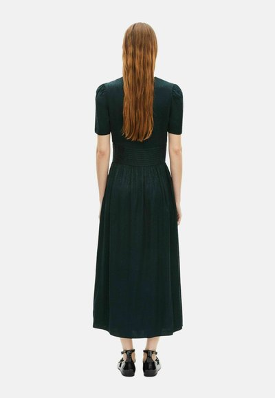 Maxi-Kleid aus dunkelgrünem Stoff mit kurzen Puffärmeln, einem gerafften Bund und einem fließenden Rock. Getragen mit schwarzen Sandalen.