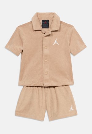 Completo beige con camicia a maniche corte abbottonata e pantaloncini coordinati con logo Jumpman bianco sul petto e sulla gamba sinistra.