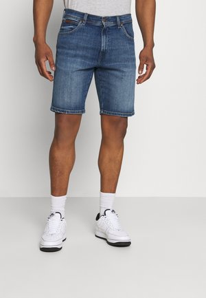 Uomo con pantaloncini di jeans blu di lunghezza media, calzini bianchi e scarpe da ginnastica bianche, in piedi davanti a uno sfondo chiaro e semplice.