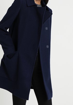 Kobieta ubrana w ciemnogranatowy płaszcz z dużymi guzikami i bocznymi kieszeniami, w połączeniu z czarnymi spodniami na jasnym, jednolitym tle.