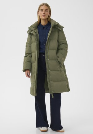 Olivgrüner gesteppter Mantel mit Kapuze, Druckknöpfen und Kordelzug am Saum; kombiniert mit einem dunklen Denim-Hemd und weit ausgestellten blauen Hosen.