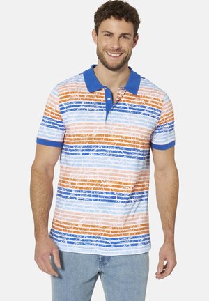 Lächelnder Mann, der ein blaues Poloshirt mit orangefarbenen, blauen und weißen horizontalen Streifen und hellblaue Jeans trägt und vor einem weißen Hintergrund steht.