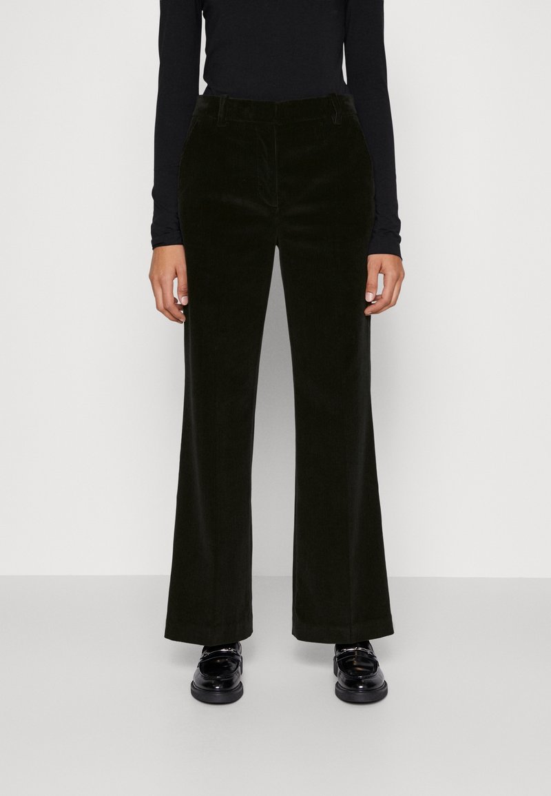 Personne portant un pantalon large en velours côtelé noir, un haut à manches longues noir et des mocassins noirs brillants, debout devant un fond blanc uni.