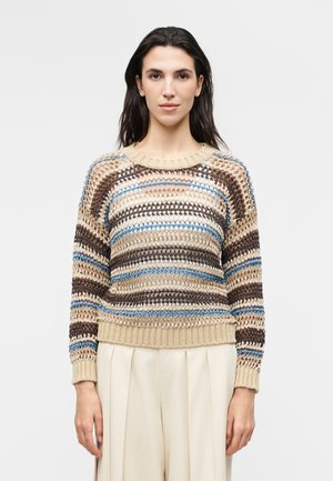 Donna che indossa un maglione a righe a maglia larga nei colori beige, marrone, blu e bianco abbinato a pantaloni crema a pieghe a vita alta su sfondo uniforme.