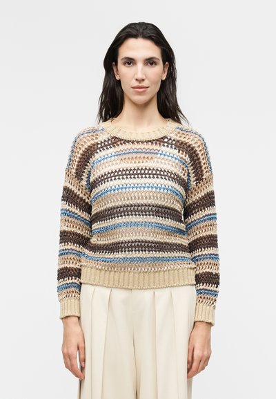 Femme portant un pull à grosse maille rayé beige, marron, bleu et blanc, associé à un pantalon plissé crème taille haute, sur un fond uni.