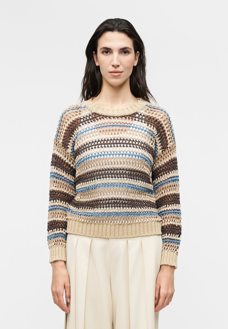 Γυναίκα με πουλόβερ σε λευκό, καφέ, μπλε και μπεζ με ριγέ μοτίβο και φαρδιά πλεκτή ύφανση, συνδυασμένο με μπεζ πλισέ παντελόνι ψηλής μέσης, μπροστά σε απλό φόντο.