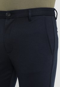 Mörkblåbyxor med en texturerad tyg, som har en framficka, bälteshällor och en enkel knäppning vid midjan.