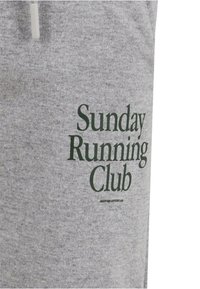 Pelēks kokvilnas sporta krekls ar lielu tumši zaļu uzrakstu "Svētdienas skriešanas klubs" priekšpusē, izceļoties ar mīkstu tekstūru un ikdienišķu dizainu.