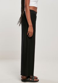 Pantalons larges noirs à côtes avec une taille élastique confortable, associés à des sandales noires, vus de côté. Texture lisse.