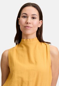 Mosterdgeel mouwloos topje met een hoge halslijn, gemaakt van lichte stof, met een gladde textuur en subtiele stikdetails.
