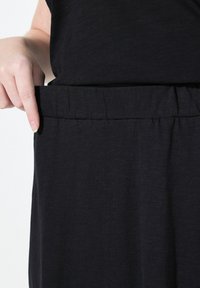 Sorte kjole med en blød tekstur, forsynet med et elastisk liv. En hånd vises, mens den holder stoffet ved hofteområdet.