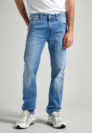 Jeans de denim azul claro con un corte recto, cinco bolsillos, un ligero desvanecimiento y cierre de botón. Usados con zapatillas deportivas blancas y grises.