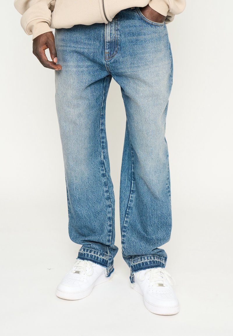 Homme portant un jean en denim bleu clair avec des ourlets bruts et des baskets blanches, debout avec une main dans la poche.