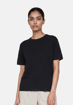 Jeune femme aux cheveux foncés attachés en arrière porte un t-shirt noir uni à manches courtes et un pantalon clair sur fond blanc.