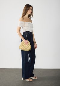 Beige off-shoulder top, dark blue high-waisted jeans, and a textured yellow woven handbag with a chain strap. Small, strappy white heels.