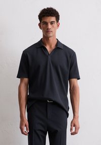 Schwarzes Polo-Shirt mit kurzen Ärmeln aus strukturiertem, geripptem Stoff, mit Ausschnitt-Kragen und lockerer Passform. Kombiniert mit dunklen Hosen.