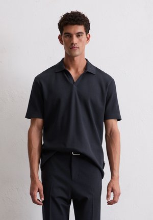 Black short-sleeve polo shirt with a textured, ribbed fabric, featuring a notched collar and a relaxed fit. Paired with dark trousers.
