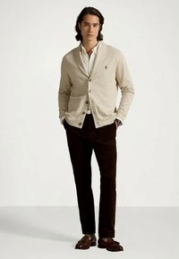 Mężczyzna w beżowym kardiganie na białej koszuli, ciemnych spodniach i brązowych mokasynach, stojący z rękami w kieszeniach na jednolitym tle.