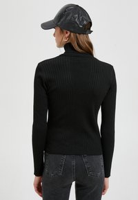 Schwarzer gerippter Rollkragenpullover mit langen Ärmeln, kombiniert mit einer schwarzen Lederbaseballkappe. Der Stoff ist strukturiert und figurbetont.