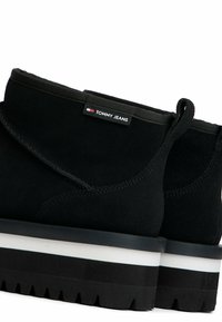 Botas de tobillo de ante negro con ribete negro, que presentan una suela gruesa blanca y negra, y una pequeña etiqueta "TOMMY JEANS" en el lado.