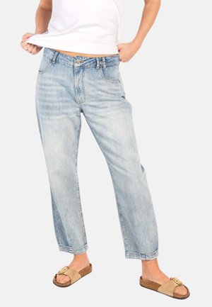 Person som har på seg lys blå jeans med rette ben, beige sandaler med åpen tå og en hvit skjorte løftet litt for å vise livet.