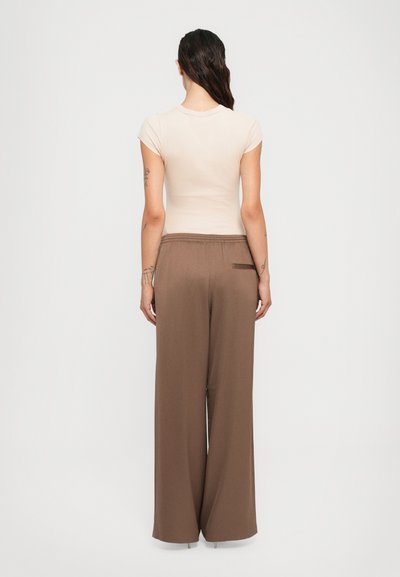 Woman with long dark hair in a low ponytail wears a cream short-sleeve top and high-waisted wide-leg brown pants, standing facing away.