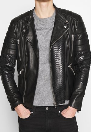 Homme portant une veste de motard en cuir noir avec des panneaux texturés à motif serpent et des fermetures éclair argentées sur un t-shirt gris à col rond.