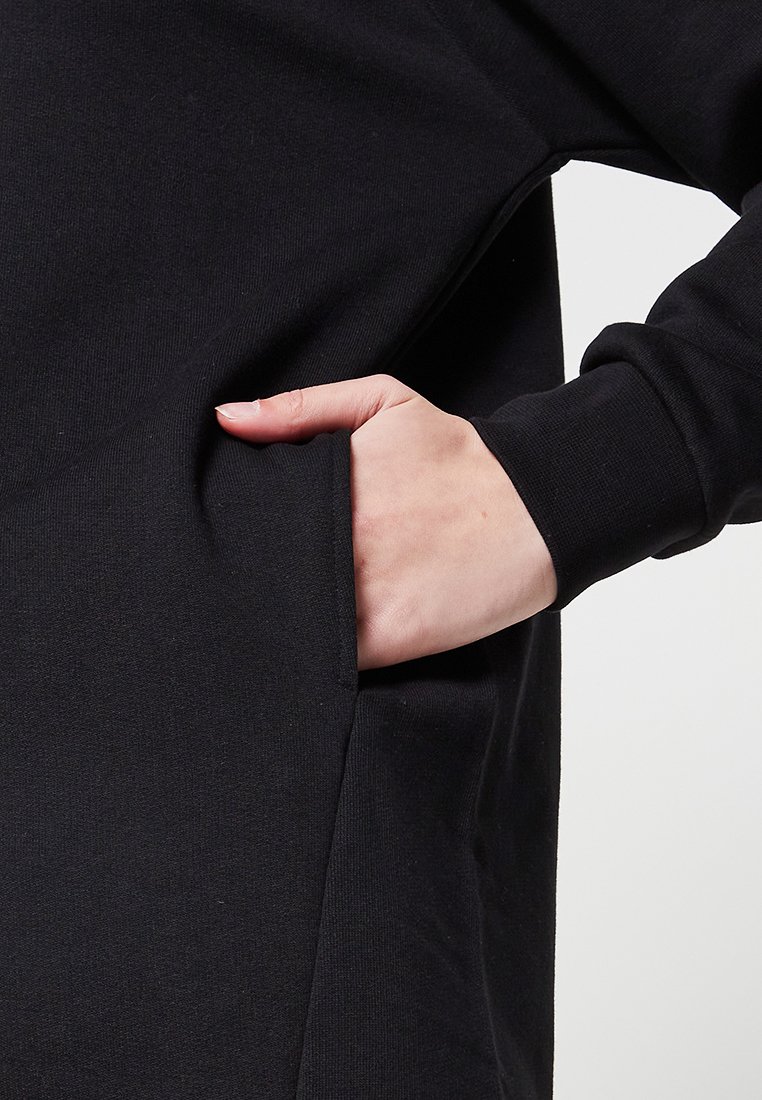 Czarny sweatshirt z boczną kieszenią, widoczną ręką spoczywającą w środku, wykonany z miękkiego, teksturowanego materiału o luźnym kroju.