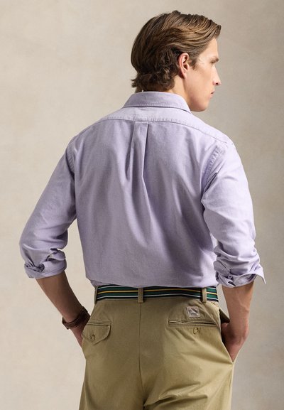 Mężczyzna stojący tyłem, ubrany w jasnofioletową koszulę z długim rękawem podwiniętym do łokci oraz beżowe spodnie z paskiem w paski.