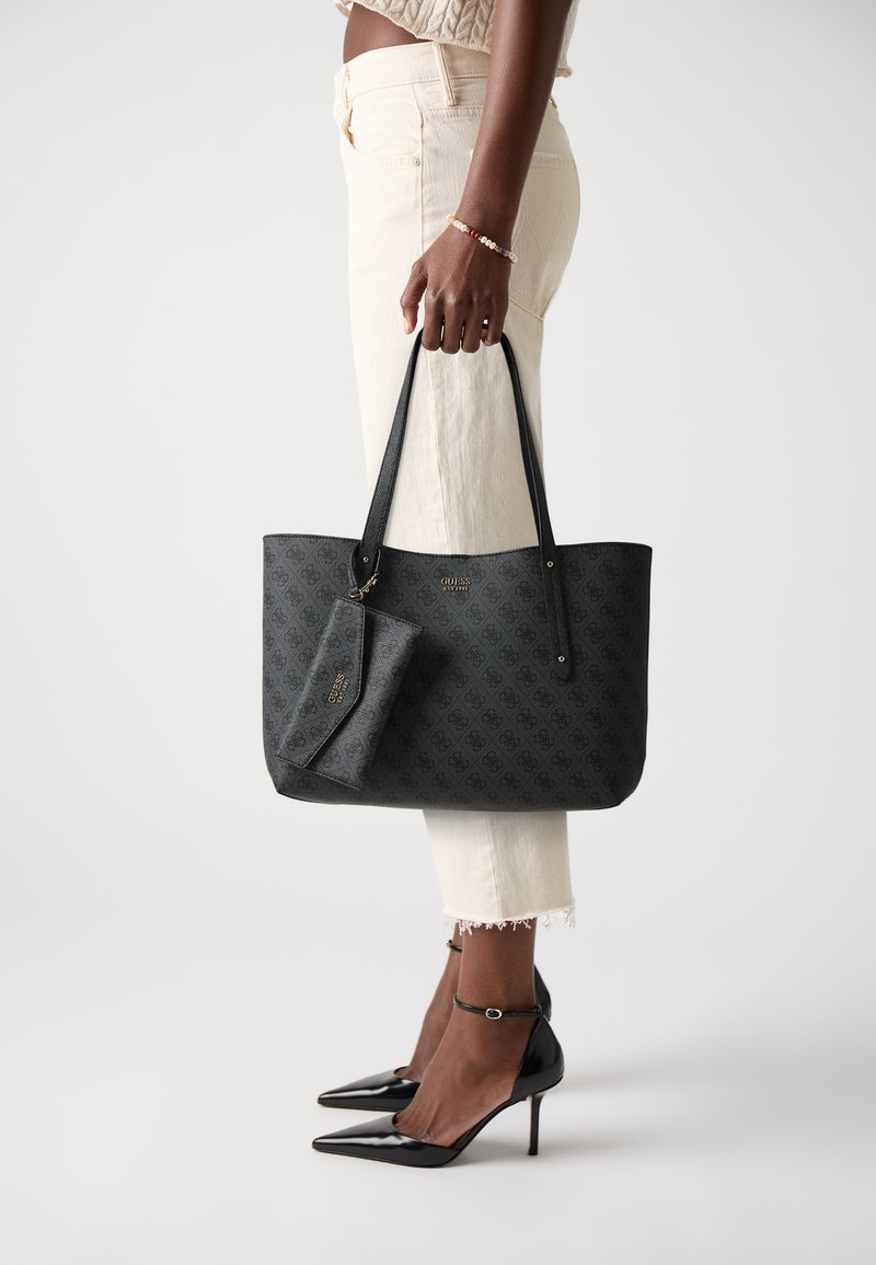 Schwarze gemusterte Tote-Bag mit langen Griffen, ausgestattet mit einem abnehmbaren kleinen Täschchen. Tragen mit hellen Hosen und schwarzen spitzen Absätzen.