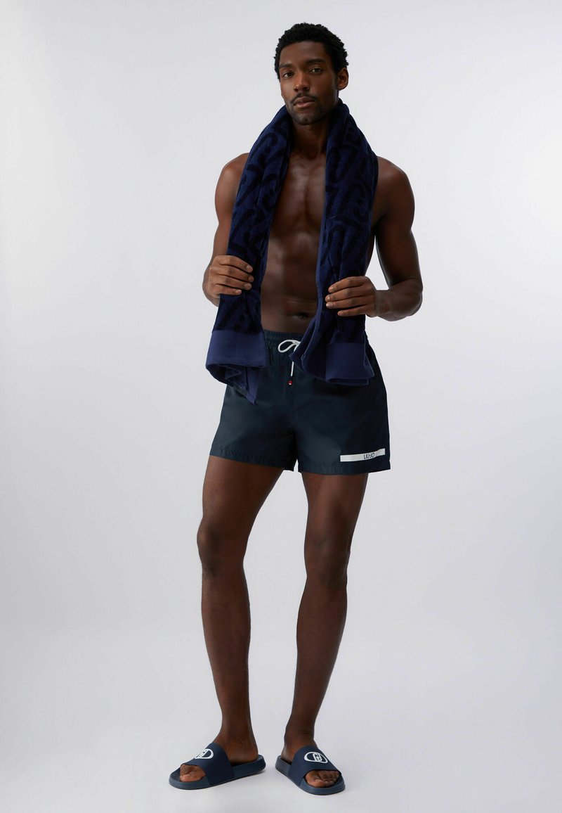Homme portant un short de bain bleu marine et des claquettes assorties, tenant une serviette bleu foncé autour du cou, debout devant un fond clair uni.