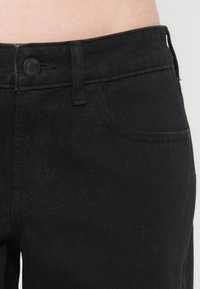 Κοντινό πλάνο στη μέση μαύρου τζιν με κλείσιμο κουμπιού, θηλιά ζώνης και λεπτομέρεια μπροστινής τσέπης σε ανοιχτόχρωμο δέρμα.