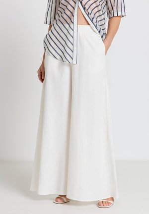 Donna che indossa pantaloni bianchi a gamba larga e vita alta, camicia a righe infilata parzialmente con bottoni e sandali bianchi aperti, in piedi contro uno sfondo neutro.