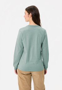 Langarmshirt aus weichem, gedämpftem grünem Baumwollstoff. Verfügt über einen runden Halsausschnitt und eine lockere Passform, kombiniert mit beigen Cordhosen.