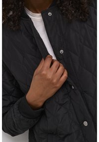 Schwarze gesteppte Jacke mit Druckknöpfen, gerippten Bündchen und glatter Textur. Eine Hand öffnet die Jacke und zeigt ein weißes Hemd darunter.