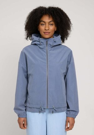 Woman with curly hair wearing a blue zip-up hooded jacket with drawstrings and light blue pants, facing forward against a plain background.