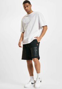 Fehér pamut póló, amelyhez fekete sportshorts tartozik, nagyméretű szövegnyomattal. Fehér sportcipővel és látható bokazoknival viselve.