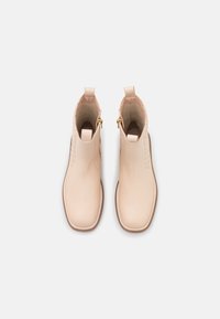 Botines de piel beige hasta el tobillo con un acabado suave, punta redonda y panel trasero texturizado. Cuentan con lengüetas y cremalleras laterales para facilitar su uso.