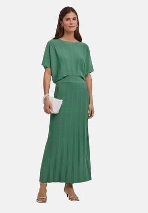 Vrouw draagt een groene geribbelde gebreide jurk met korte mouwen en een lange rok, houdt een witte clutch vast en draagt open-toe hakken.