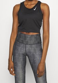 Schwarzes, kurz geschnittenes sportliches Tanktop mit weißem Nike-Swoosh-Logo. Kombiniert mit eng anliegenden schwarzen Leggings, die ein sich wiederholendes weißes "Nike"-Muster aufweisen.