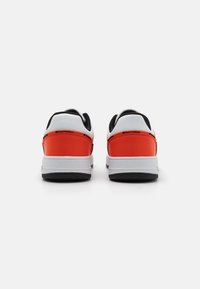 Vue arrière des baskets blanches et orange avec des accents noirs, talon texturé et semelle en caoutchouc plate. Présente un branding minimal près du talon.
