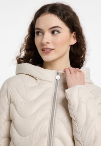 Beige gewatteerde pufferjack met een capuchon, voorzien van golvende stiksel, een zilveren rits en een subtiele glans. Zacht buitenmateriaal met een gladde textuur.