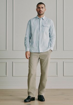 Mann in beiger Hose, hell gestreiftem Hemd mit Knöpfen und schwarzen Schuhen steht vor einer grauen, vertäfelten Wand.