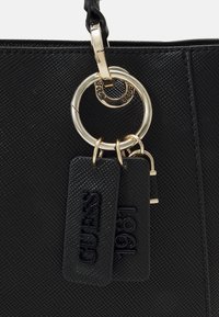Sort tekstureret Guess-håndtaske med guld ringe og to sorte mærker præget med "GUESS" og "1981".